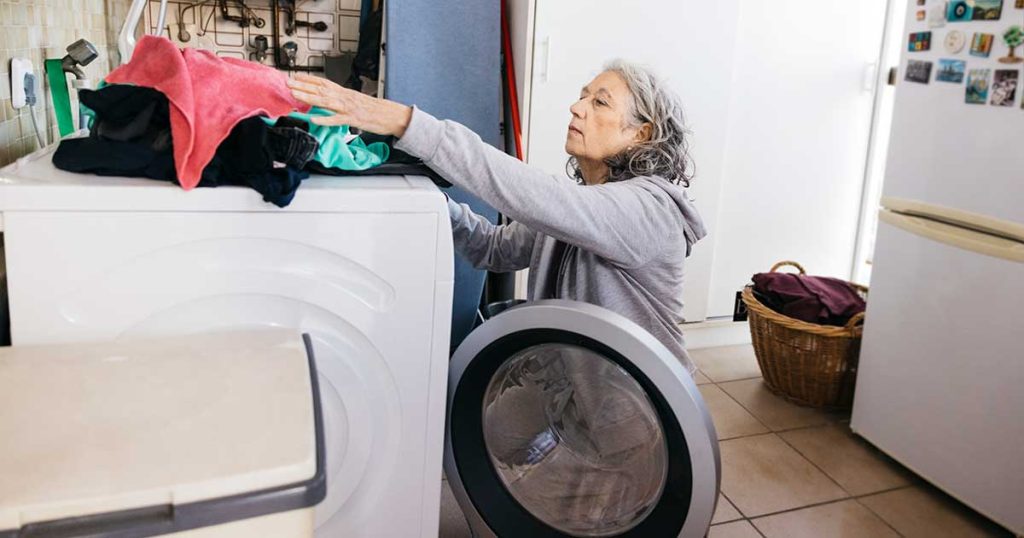 A woman with wavy, light grey hair, wearing a grey sweatshirt, is putting clothes in a white washing machine