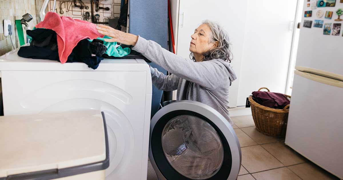 A woman with wavy, light grey hair, wearing a grey sweatshirt, is putting clothes in a white washing machine